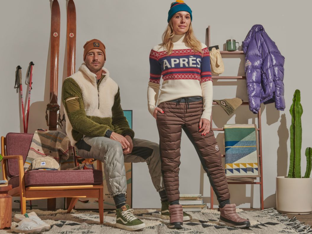 A man and woman wearing down sweatpants and retro-inspired ski sweaters lounge in a cozy après-ski setting.